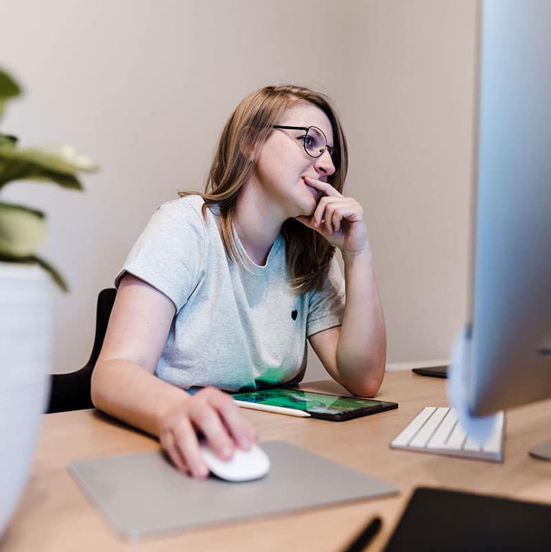 Fokussiert und lösungsorientiert – Webdesign mit Struktur Tine, Webdesignerin, sitzt am Schreibtisch vor einem großen Monitor und schaut nachdenklich auf den Bildschirm. Ihre Hand liegt an der Maus, während sie sich auf das Design oder die technische Umsetzung konzentriert.